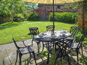 An outdoor patio with a round table set for dining, surrounded by metal chairs, in a garden with a brick wall and greenery in the background.