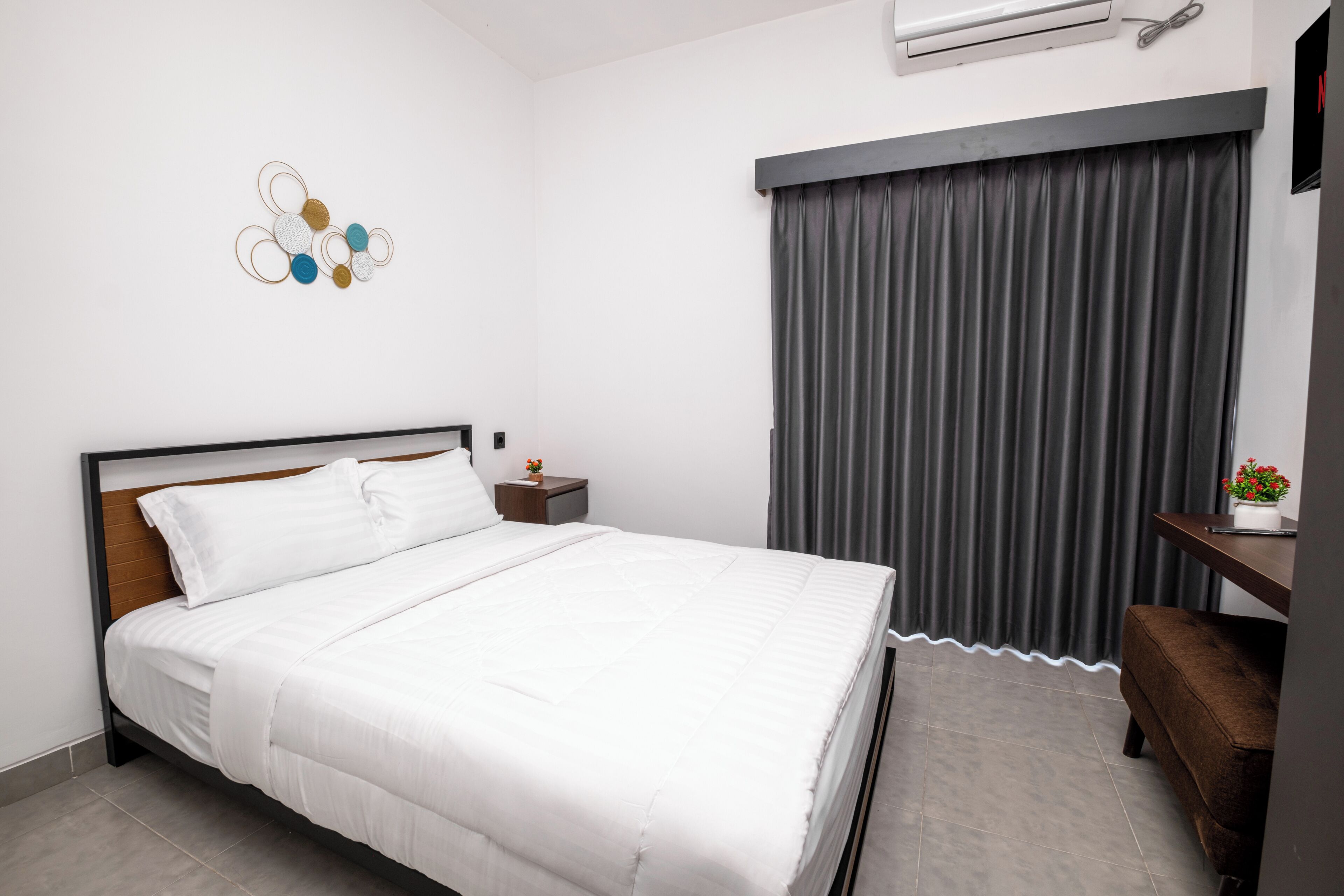 A neatly made bed in a hotel room with a headboard, a bedside table with a lamp, a wall-mounted air conditioner, and a window with curtains.