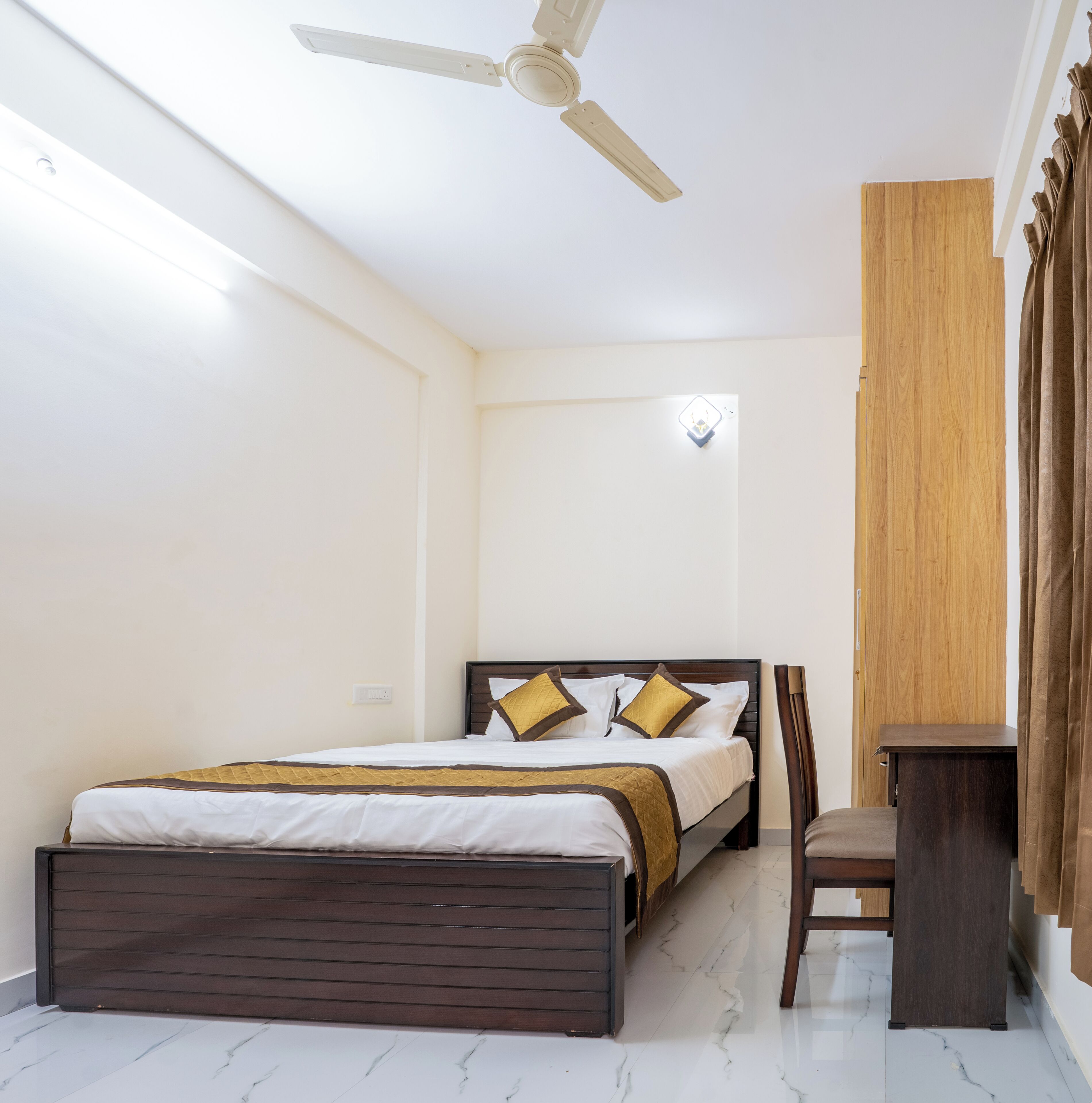 Une chambre à coucher comprenant un lit, une chaise, un bureau, un ventilateur de plafond et une armoire en bois.