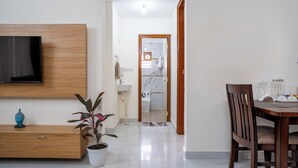 A modern living room with a TV, wooden entertainment unit, potted plants, a dining area with a table and chairs, and a hallway leading to a bathroom. - Odilia Apartelle (Bengaluru)