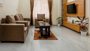 A modern living room with a sofa set, a coffee table, a TV mounted on a wooden panel, a ceiling fan, and a window with curtains. - Odilia Apartelle (Bengaluru)