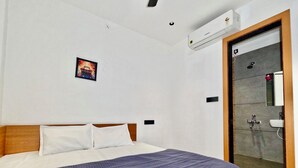 Ein Hotelzimmer mit einem Bett, einer Wandklimaanlage, einem kleinen Bild und einem durch eine Glastür einsehbaren Badezimmer.