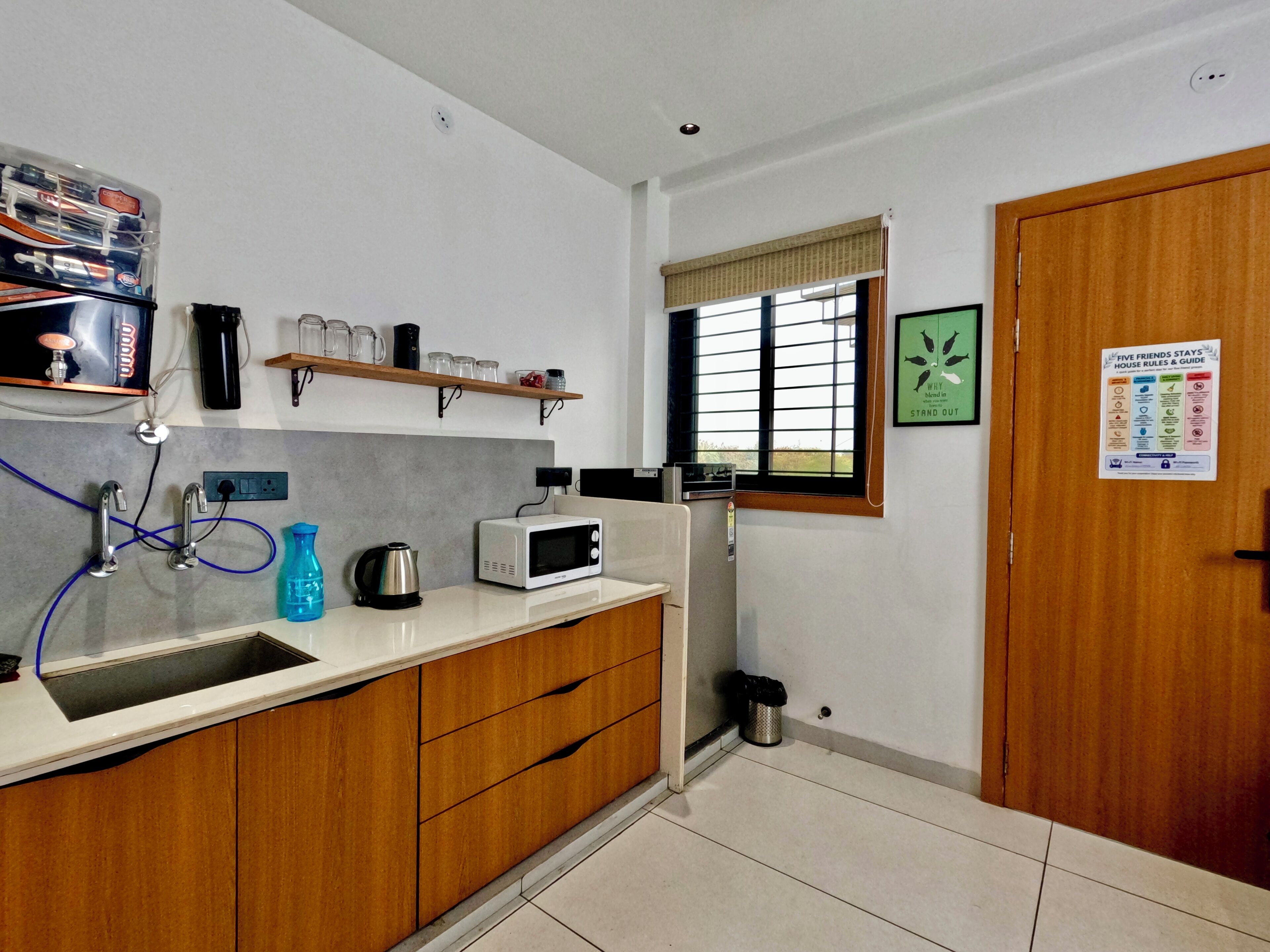 A kitchen with a microwave, kettle, and sink; a window with blinds; and a door with a house rules poster.
