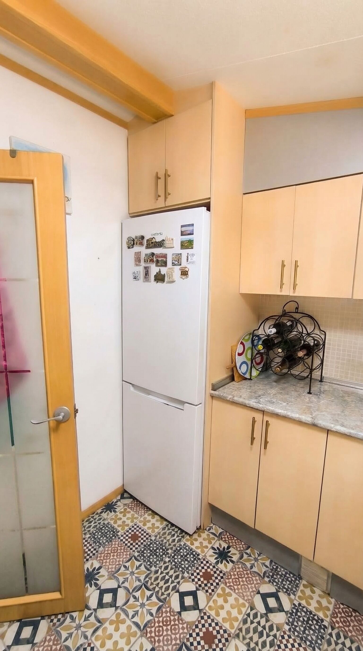 A compact kitchen with a refrigerator, wooden cabinets, a wine rack, and patterned floor tiles.