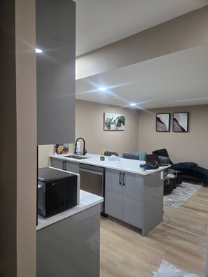 A compact kitchen with a microwave, sink, and cabinets, a small seating area with a couch and a coffee table, and two framed artworks on the wall. - "Your Home Away From Home" (Edmonton)