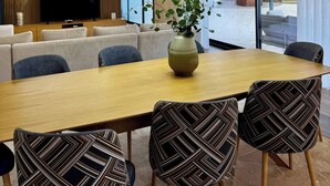 Moderni ruokailutila, jossa on pitkä pöytä, kuviollisia tuoleja, maljakko, jossa on kasveja, ja taustalla näkyy televisio.