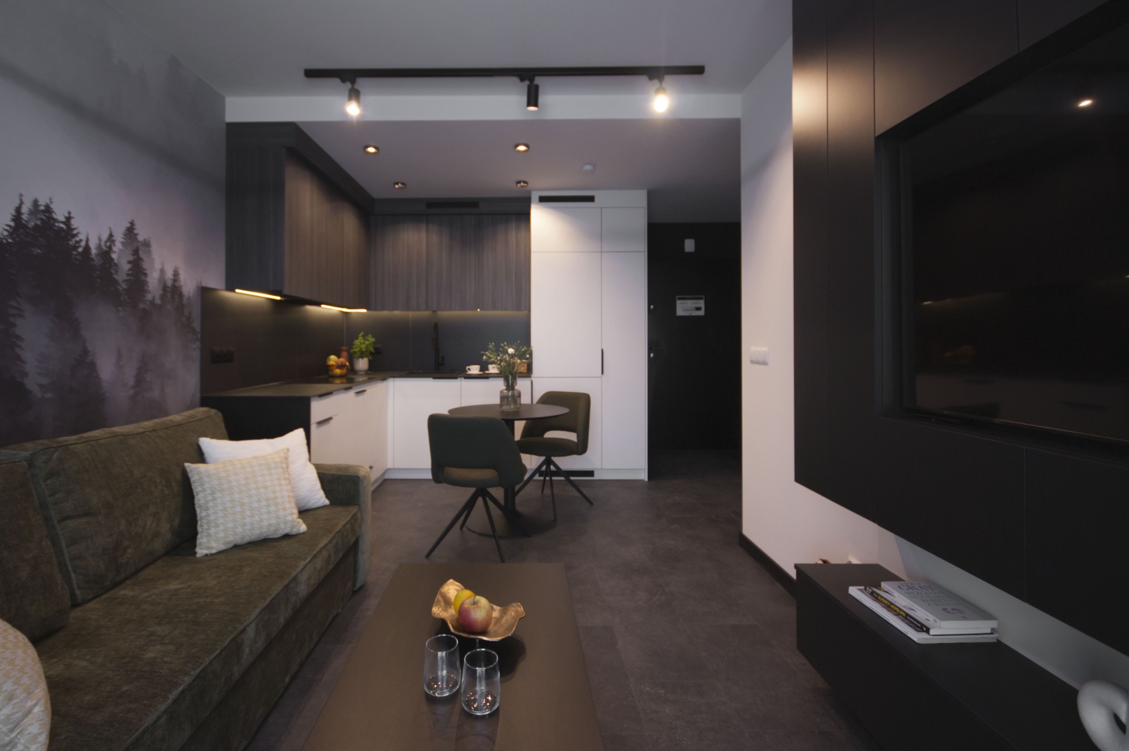 Una sala de estar moderna con un sofá, una mesa de centro con frutas y vasos, un televisor, y una zona de cocina con una mesa de comedor y sillas.