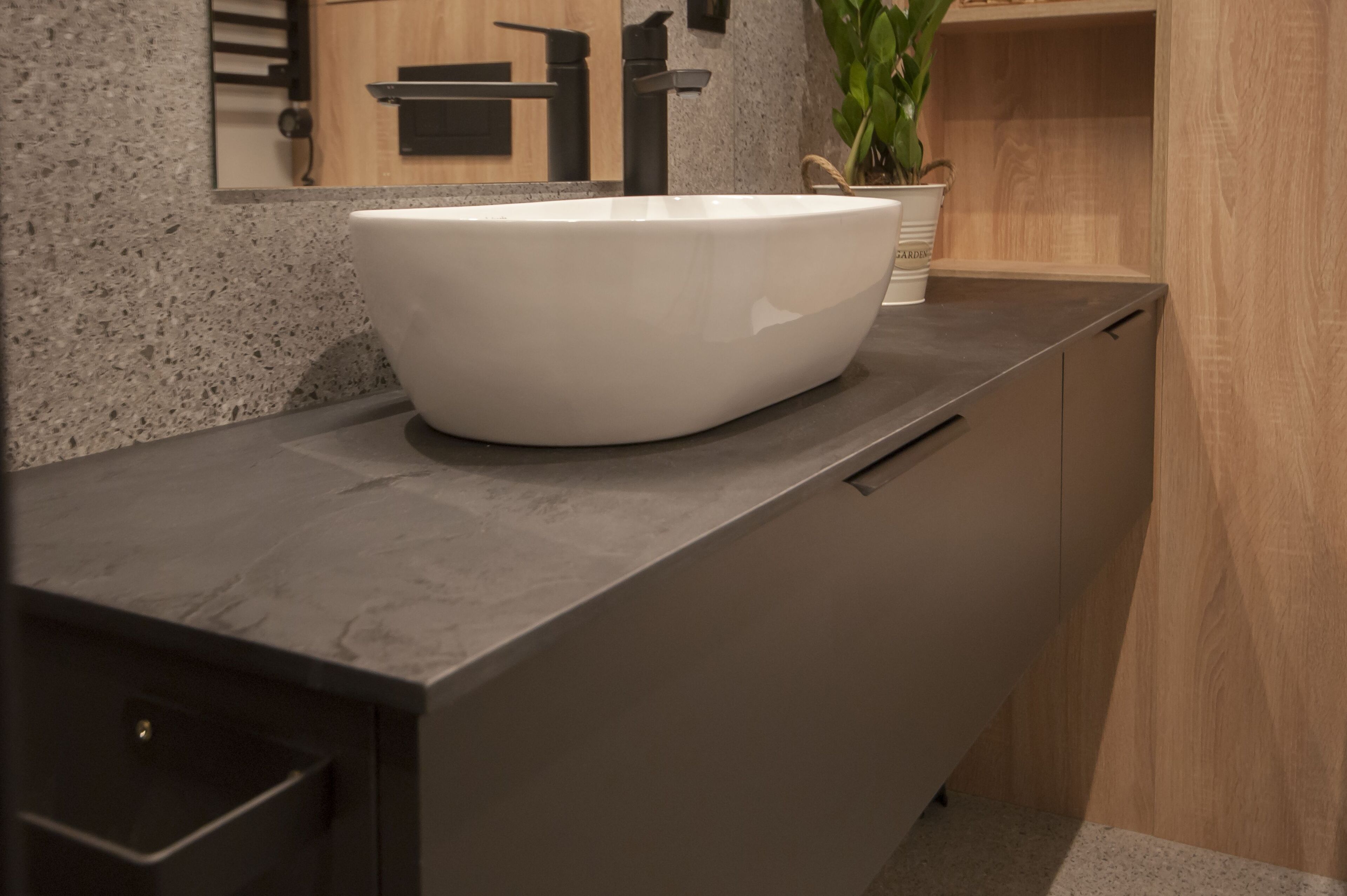 Un baño moderno con un lavamanos blanco de diseño, una encimera oscura y un mueble de madera.