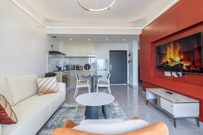 Una sala moderna con un sofá, una mesa de centro redonda, un televisor con una pantalla de chimenea y una cocina al fondo.