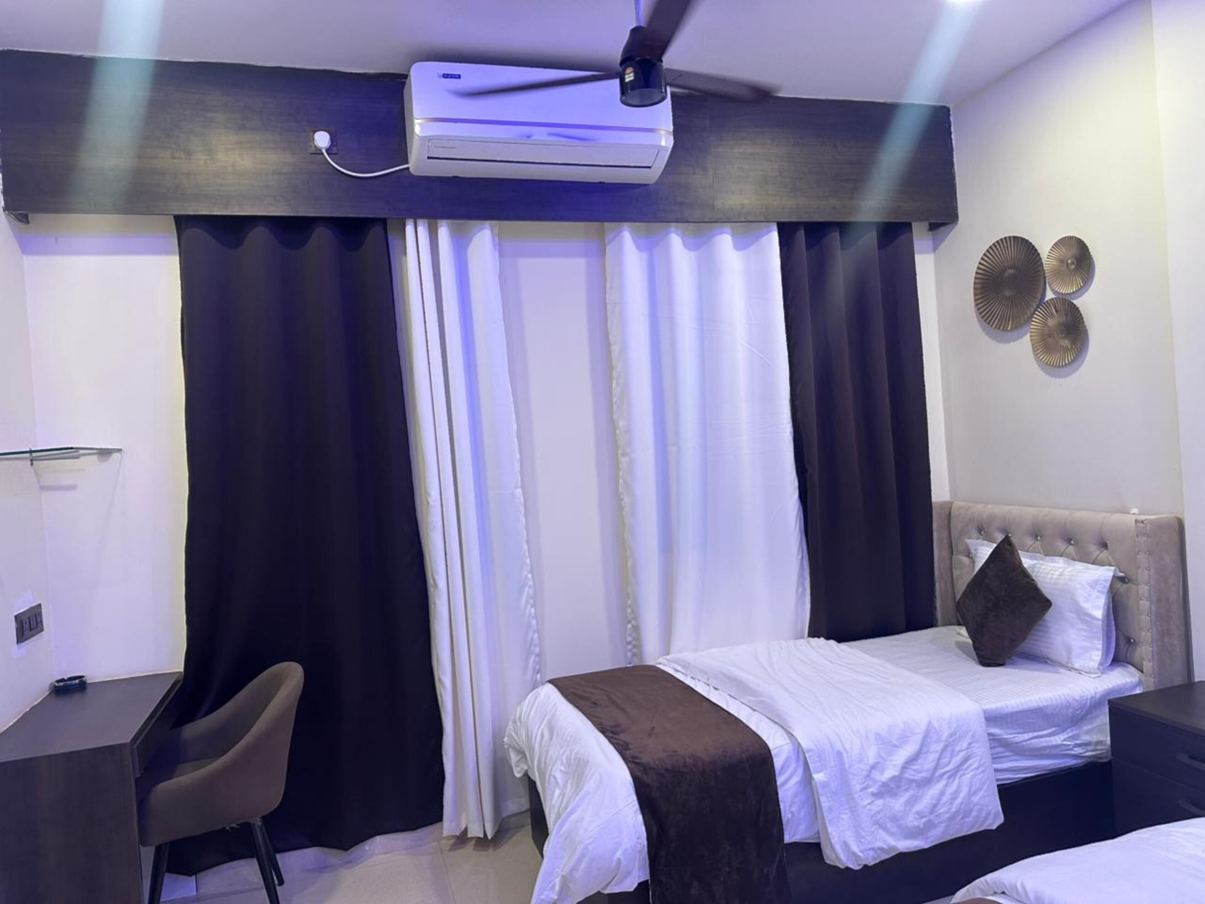 Une chambre d’hôtel équipée d’un lit, d’un bureau, d’une chaise, d’un ventilateur de plafond et d’une fenêtre avec des rideaux.