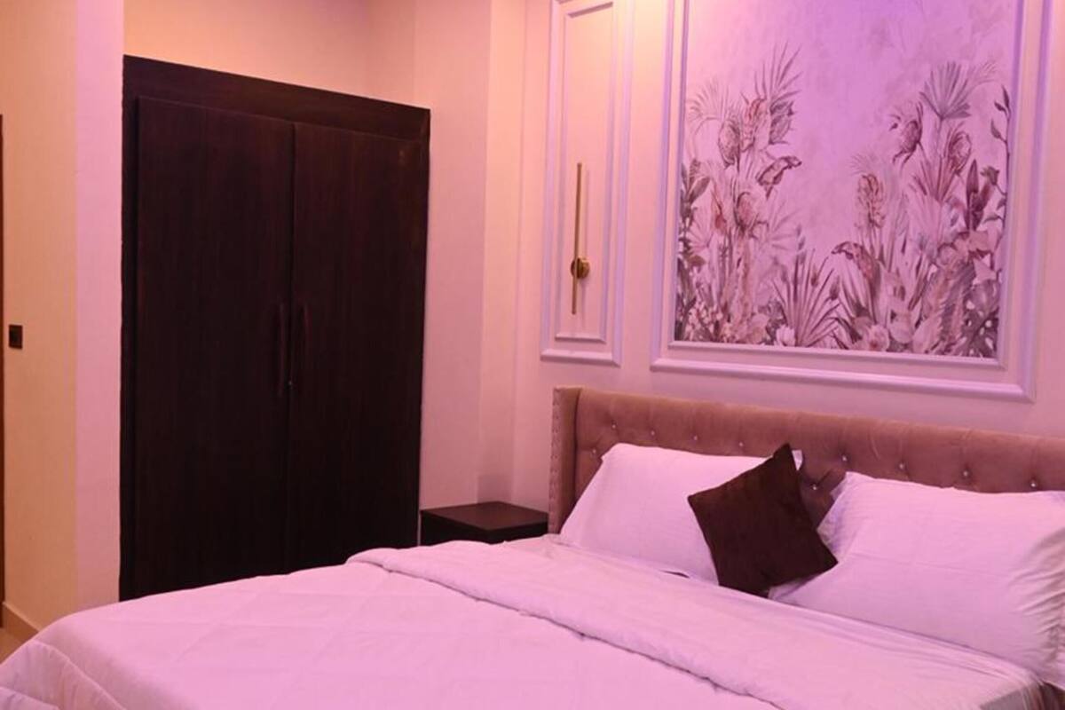 A bedroom with a bed, a ceiling fan, a wardrobe, and a floral painting on the wall.
