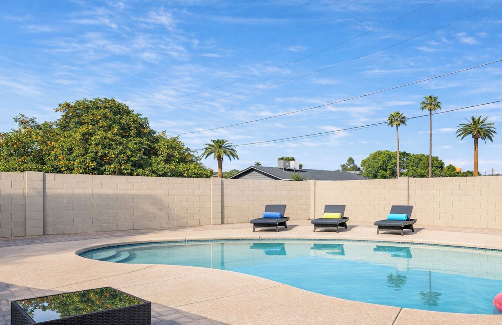 A swimming pool with three lounge chairs, a brick wall, and palm trees in the background.