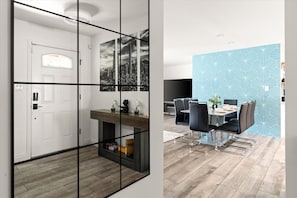 Ein modernes Interieur mit Holzboden, einer weißen Tür, einem Spiegel mit schwarzem Rahmen, einem Essbereich mit Tisch und Stühlen und einer Wand mit blauem geometrischen Muster.