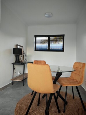 Een eetgedeelte met een ronde glazen tafel, twee oranje stoelen, een zwarte lamp en een raam met jaloezieën.