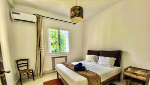 Una camera d'albergo con un letto, una sedia, una finestra con tende, un condizionatore d'aria e un comodino in legno.