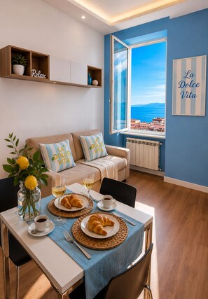 Un salon moderne avec un canapé, une table à manger dressée avec des croissants, du café et du vin, et une vue sur la mer par une grande fenêtre.