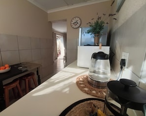 Una cocina con encimera blanca, cafetera negra, planta en maceta y un bol de naranjas.