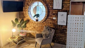 A living room with a brick wall, a round mirror, a flat-screen TV, a grey armchair, a round woven rug, and a potted plant.
