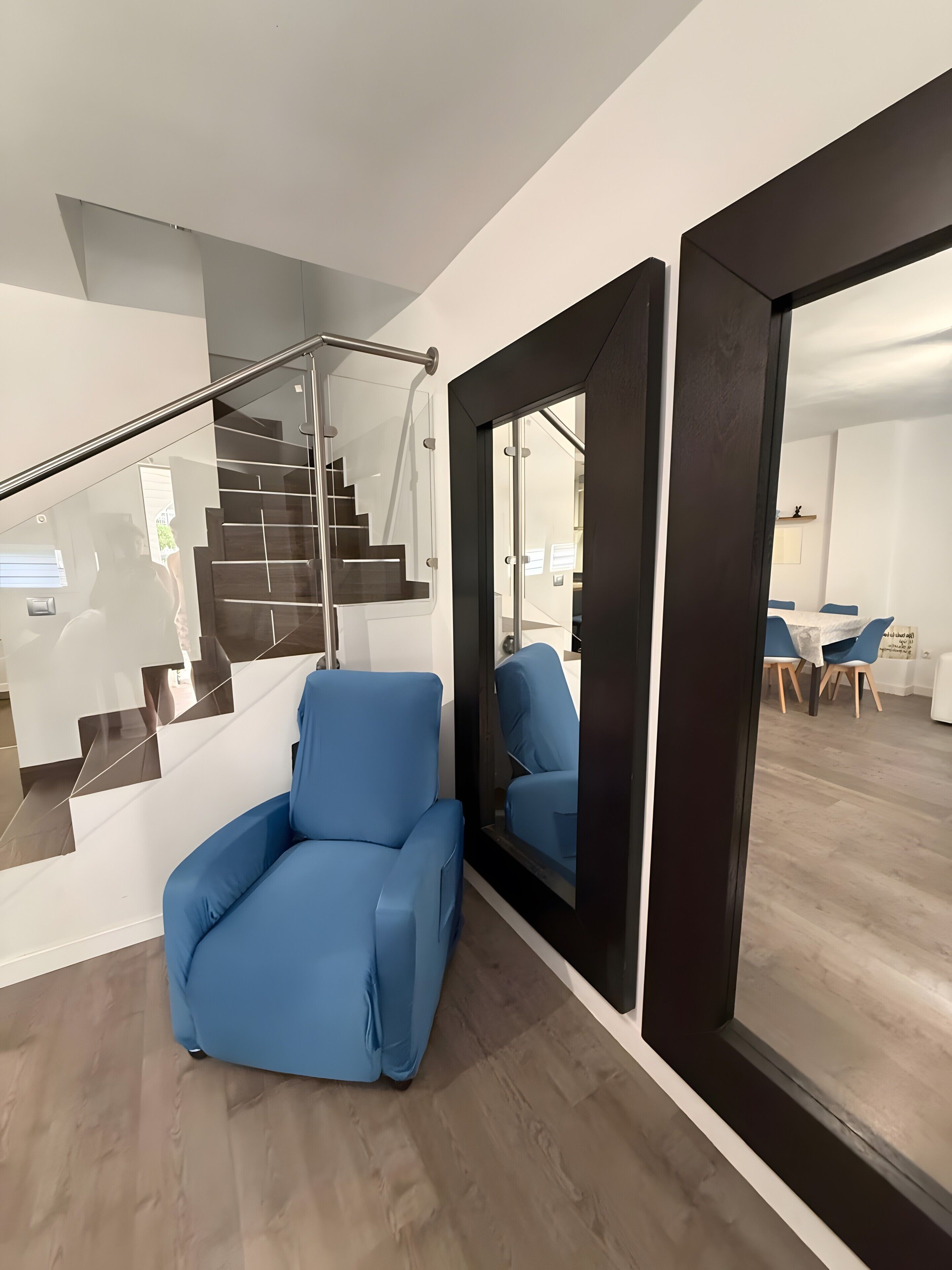 Un interior moderno con una escalera, un sillón azul y un comedor con mesa y sillas.