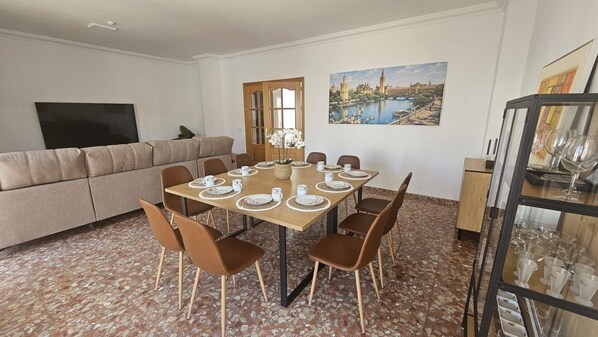 A dining area with a table set for six, a sofa, a TV, and a painting of a cityscape.