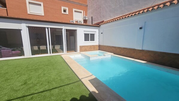 A swimming pool with a clear blue water, surrounded by a green lawn and a white building with a tiled roof.