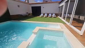 A swimming pool with a small hot tub, a grassy area with lounge chairs, and a covered patio with tables and chairs.