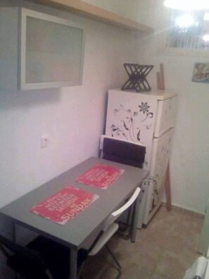 Un pequeño comedor con una mesa y sillas, un refrigerador con diseños decorativos y un estante fijado a la pared sobre la mesa.