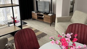 Uma sala de estar moderna com uma TV, um espaço de jantar com mesa e cadeiras, e vista através de uma grande janela.