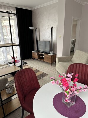 A modern living room with a TV, a dining area with a table and chairs, and a view through a large window. - Sunny Beach Central Apartment - 4 Guests, Parking (Sunny Beach)