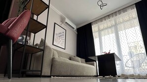 Uma sala de estar moderna com um sofá, uma poltrona vermelha, uma mesinha lateral preta, uma obra de arte emoldurada e uma janela com cortinas.