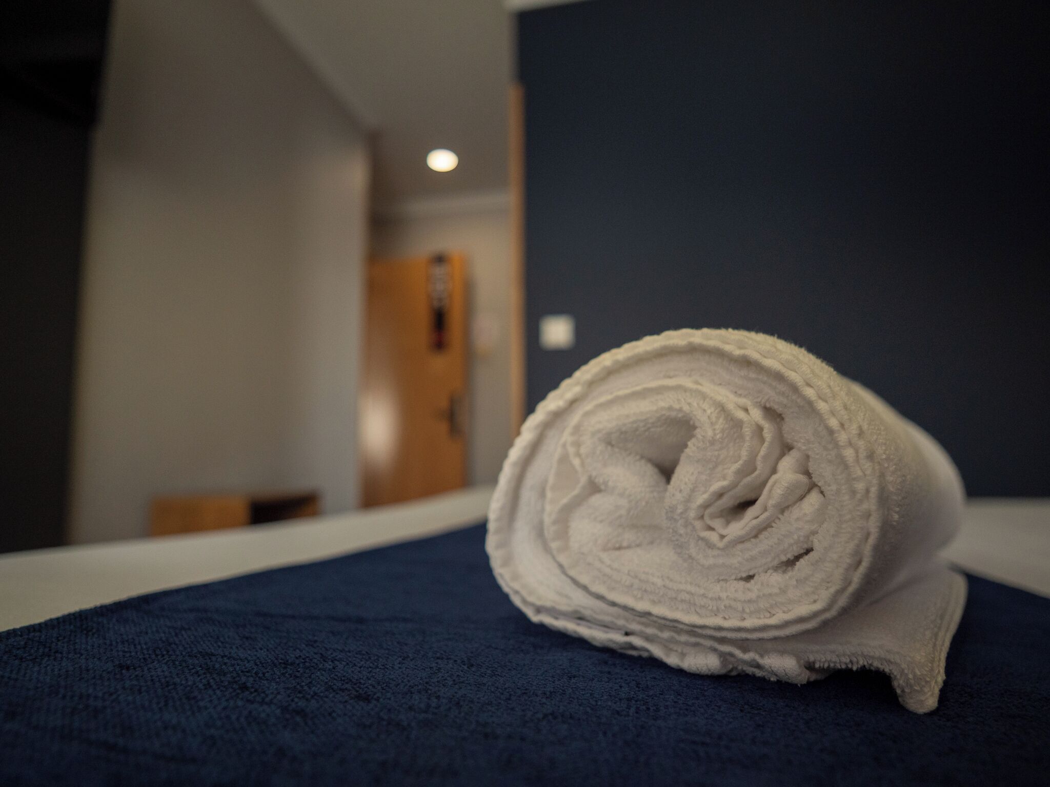 Ein gerolltes, weißes Handtuch auf einer dunkelblauen Hotelbettdecke.