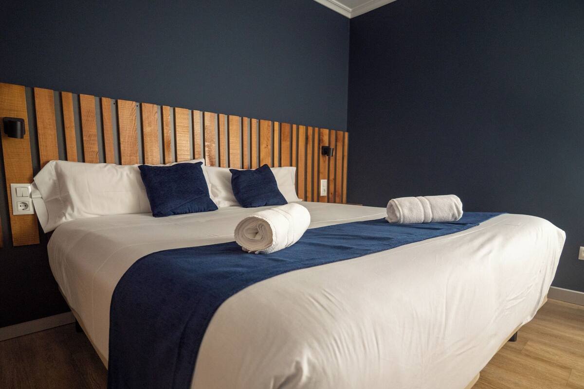 A hotel room with a wooden headboard, a large bed with white bedding, two blue pillows, and a blue throw blanket.