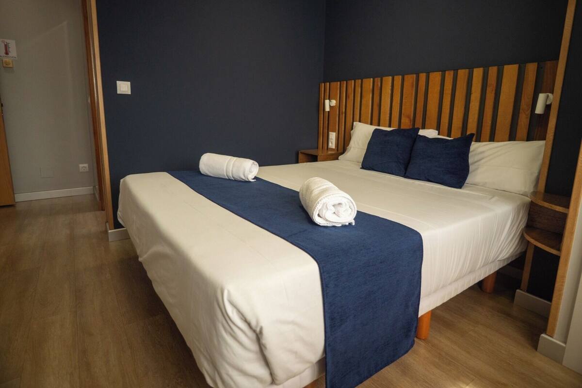 A hotel room with a bed, wooden headboard, two pillows, a blue blanket, and a rolled towel.