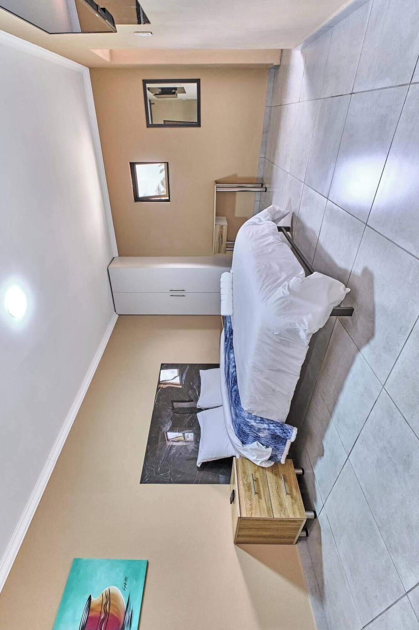 A compact hotel room with a bed, a small wooden side table, a wall-mounted towel rack, and a single framed picture on the wall.