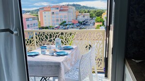 Un balcon avec une table dressée pour deux personnes, un luminaire suspendu et une vue sur un quartier résidentiel avec ses immeubles et ses voitures garées.
