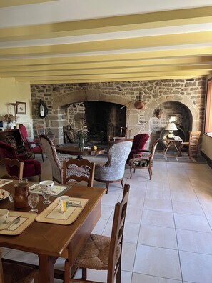 A cozy living room with a stone fireplace, wooden furniture, and a dining area with a table set for two. - Quiet vacation home with swimming pool (Bagnols)