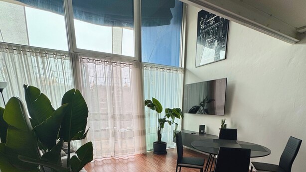 Una habitación amplia con piso de madera, una mesa de comedor con sillas, una planta en maceta, un ventanal con cortinas translúcidas y un televisor de pantalla plana montado en la pared.
