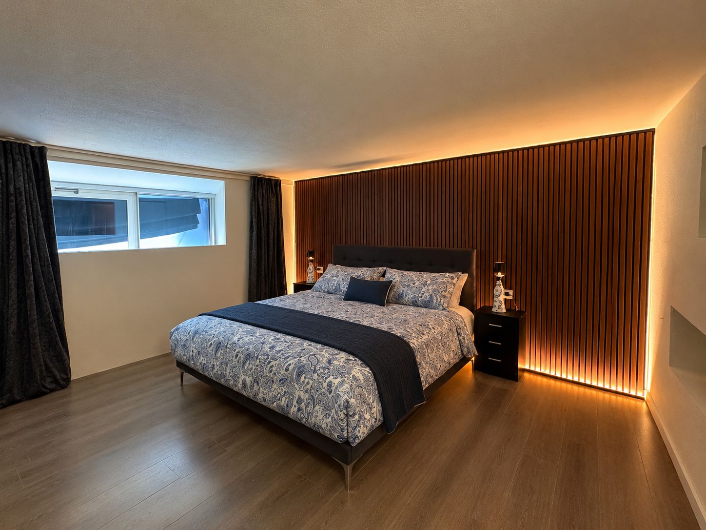 Ein modernes Schlafzimmer mit einem großen Bett, einem Nachttisch, einem Fenster und einer Wand mit vertikalen Holzleisten.