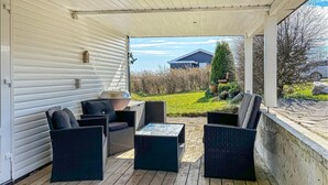 A covered patio with wicker furniture, a grill, and a view of a house and greenery.