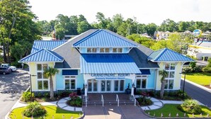 A hotel with a blue roof, a sign reading 'Grand Palms Resort,' and a landscaped entrance with American flags. - Myrtle Beach | Friends Golf Retreat | Pool + Golf | 2BR (Surfside Beach)