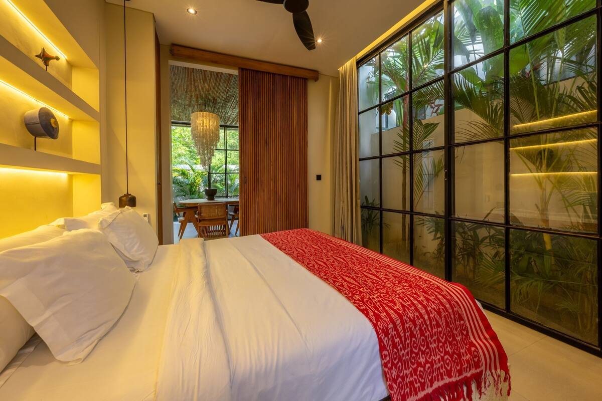 A bedroom with a bed, a dining table, and a large window with a view of greenery.