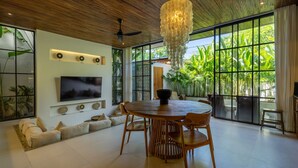 A modern dining area with a round wooden table, chairs, a large TV, and a ceiling fan. - Aricivillas (Pecatu)