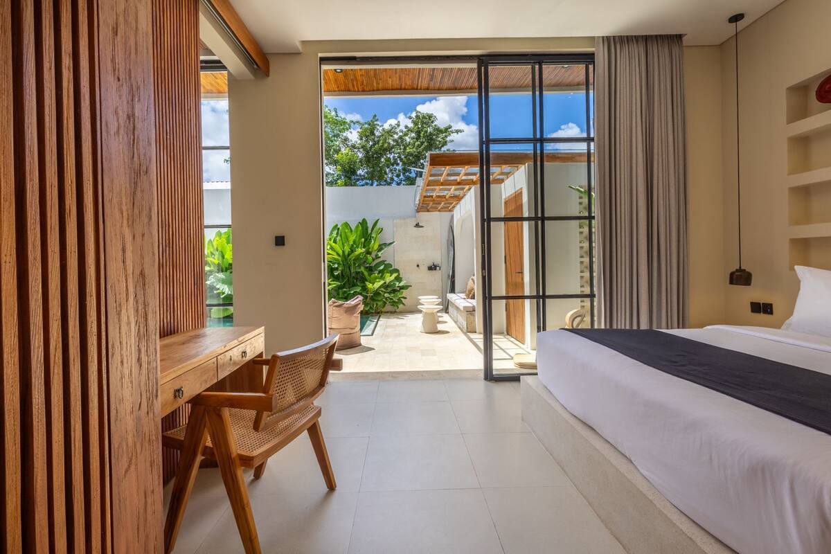 A modern hotel room with a large bed, a wooden desk, and a view of the outdoor garden.