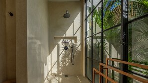 A small, sunlit room with a shower, a wooden stool, and a window with a view of greenery. - Aricivillas (Pecatu)