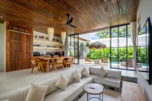 A modern interior with a wooden ceiling, a dining area, a sofa, and large windows overlooking a garden. - Aricivillas (Pecatu)