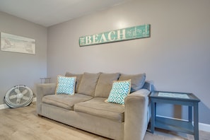 Ein beiges Sofa mit gemusterten Kissen, ein kleiner Beistelltisch, ein an der Wand befestigtes Schild mit der Aufschrift 'THE BEACH' und ein Ventilator an der Wand.
