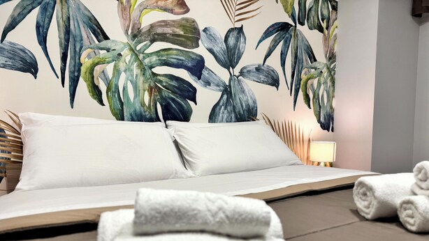 A hotel room with a bed, white pillows, a nightstand with a lamp, and a wall mural of tropical leaves. - Modern apartment in the center of Catania, private entrance, up to 6 guests WiFi (Catania)