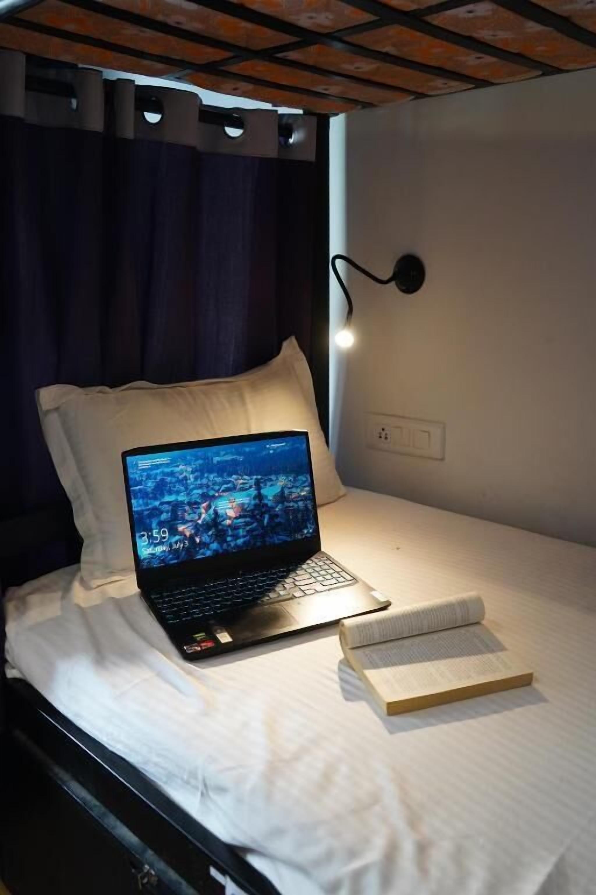 A bed with a laptop displaying a cityscape, a book, a wall-mounted light, and a power outlet.