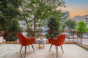 A balcony with two red chairs, a small table with a drink, and a view of buildings and trees. - Hotel AP International (Noida)