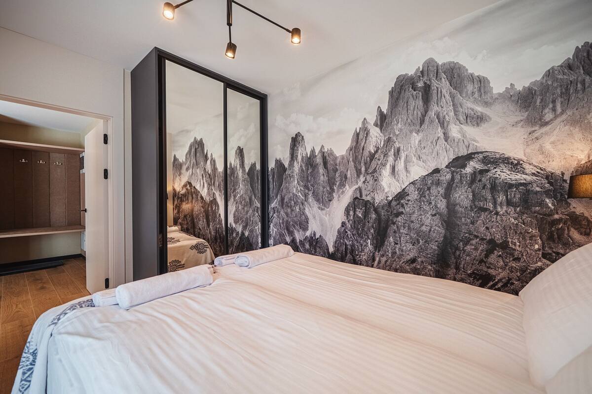 A bedroom with a large bed, a wardrobe, a mirror, and a mountain mural on the wall.